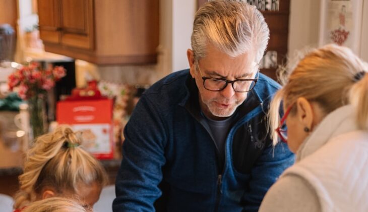 two women standing and three children sitting while preparing gingerbread house